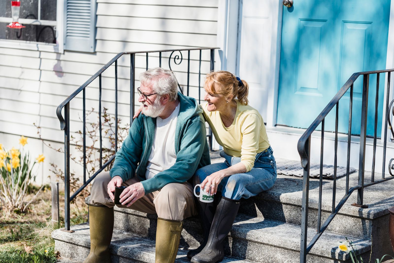 Delightful aged couple in rain boots drinking tea and looking away while sitting on stairs of house in countryside